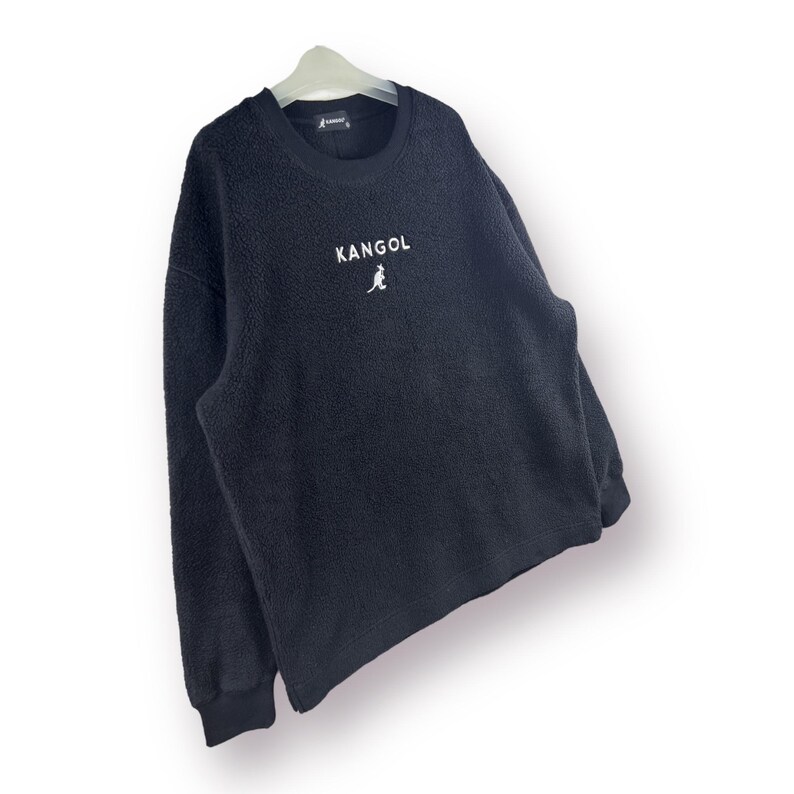 Op de afbeelding: Zwart fleece sweatshirt met een wit kangoeroe logo en het woord "Kangol" gedrukt op de voorkant.