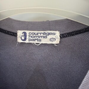 May include: Close-up of a gray crew neck shirt with a Courr&egrave;ges Homme Paris label. The label is rectangular with the brand name in white text and a blue logo. The shirt is hanging on a white hanger.