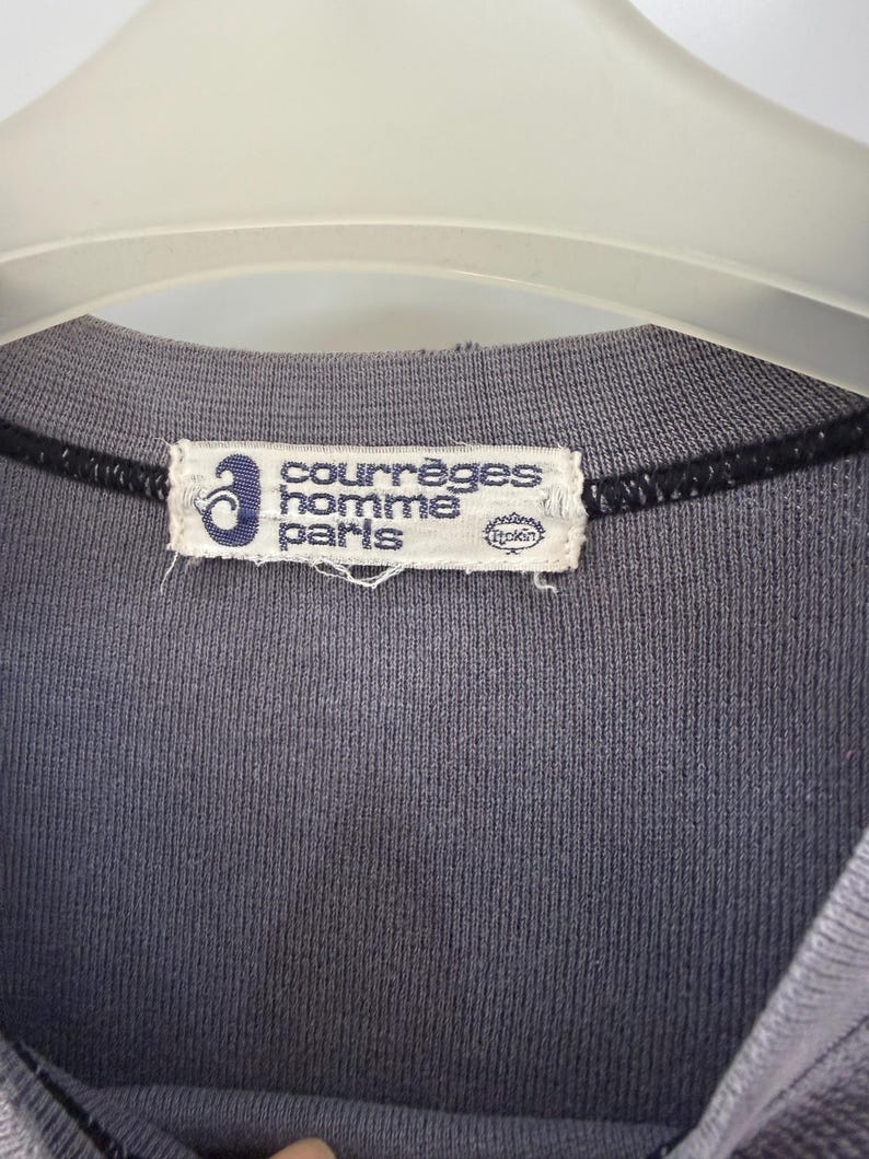 May include: Close-up of a gray crew neck shirt with a Courr&egrave;ges Homme Paris label. The label is rectangular with the brand name in white text and a blue logo. The shirt is hanging on a white hanger.