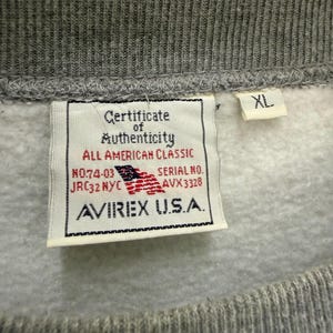 Pode incluir: Close-up de um moletom cinza com gola canelada. Uma etiqueta exibe "Certificate of Authenticity" e a marca "AVIREX U.S.A.", com uma bandeira americana. A etiqueta de tamanho diz "XL".