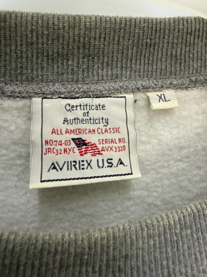 Pode incluir: Close-up de um moletom cinza com gola canelada. Uma etiqueta exibe "Certificate of Authenticity" e a marca "AVIREX U.S.A.", com uma bandeira americana. A etiqueta de tamanho diz "XL".