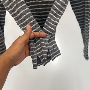 May include: A long-sleeve shirt featuring a ribbed texture with a pattern of horizontal dark gray and white stripes. The sleeves are designed with a small vertical slit at the cuff, providing a distinct detail to the garment's overall construction.