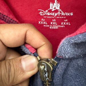 May include: Close-up of a red and navy blue zip-up hoodie with the Disney Parks logo and text. The zipper is a brass color. The hoodie has a tag with sizing information.
