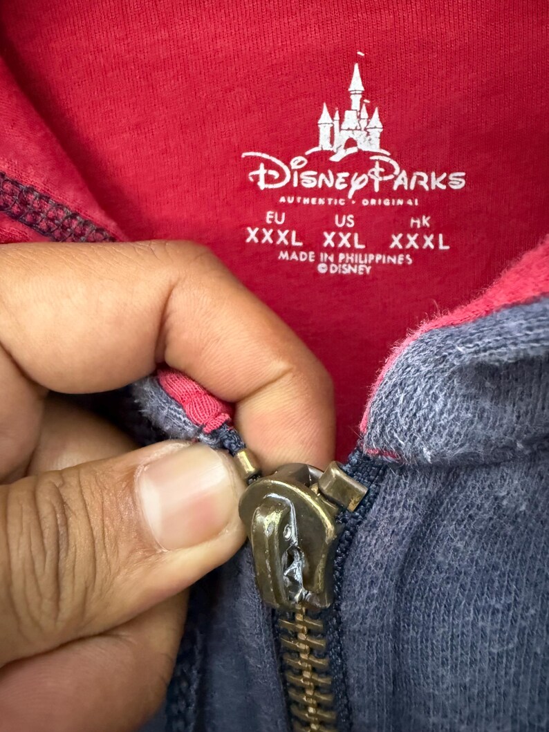 May include: Close-up of a red and navy blue zip-up hoodie with the Disney Parks logo and text. The zipper is a brass color. The hoodie has a tag with sizing information.