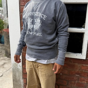 May include: A charcoal gray crewneck sweatshirt featuring a white graphic print with the text REMINISCENCE NEW YORK and two dragon illustrations. The garment has long sleeves with ribbed cuffs and a ribbed neckline with a V-shaped stitch detail.