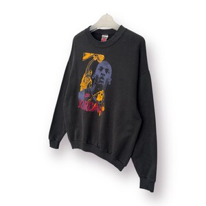 Puede incluir: Sudadera negra de cuello redondo con un gr&aacute;fico de un jugador de baloncesto en amarillo, azul y rojo. La palabra "JORDAN" est&aacute; impresa en rojo. La sudadera est&aacute; colgada de una percha blanca.