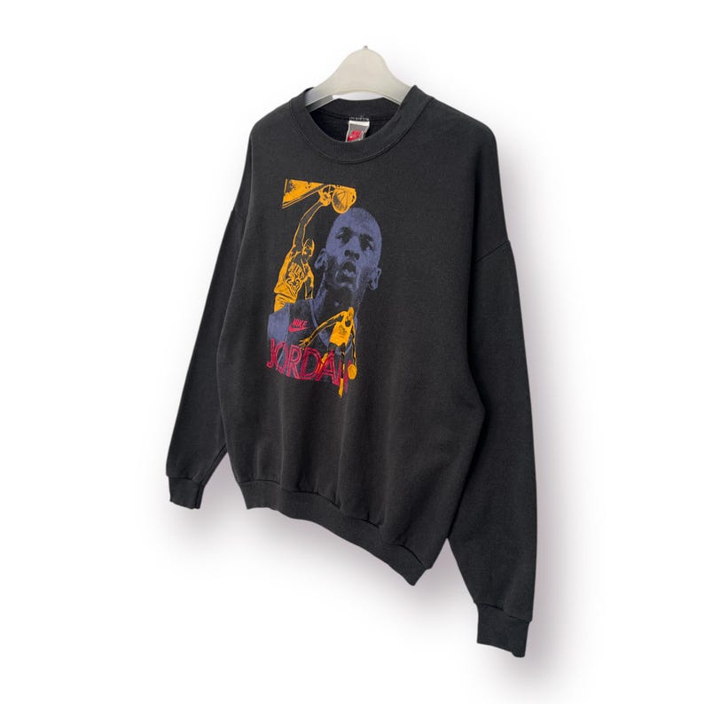 Puede incluir: Sudadera negra de cuello redondo con un gr&aacute;fico de un jugador de baloncesto en amarillo, azul y rojo. La palabra "JORDAN" est&aacute; impresa en rojo. La sudadera est&aacute; colgada de una percha blanca.