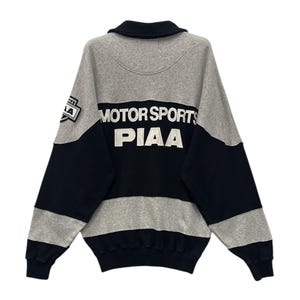 May include: A gray, black, and white sweatshirt with the text "MOTOR SPORTS PIAA" across the back. The sweatshirt has a collared neckline and a patch on the left sleeve. The sleeves and lower portion are gray.