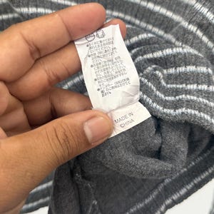 May include: A dark gray ribbed knit garment featuring horizontal light blue and white stripes. A white fabric care label is attached to the interior, displaying Japanese text instructions and the text "MADE IN CHINA" at the bottom edge.
