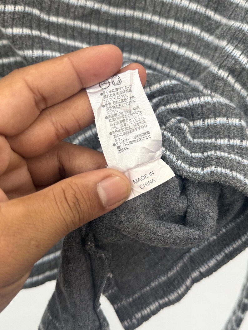 May include: A dark gray ribbed knit garment featuring horizontal light blue and white stripes. A white fabric care label is attached to the interior, displaying Japanese text instructions and the text "MADE IN CHINA" at the bottom edge.