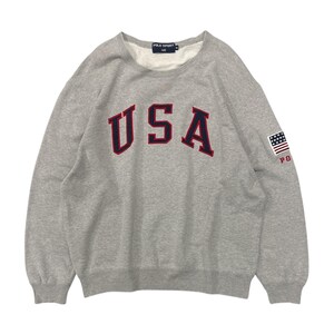 May include: A grey sweatshirt featuring the word "USA" in navy blue letters with red trim. The right sleeve has a small American flag patch and the letters "PO".