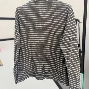 May include: A long-sleeved, ribbed knit sweater featuring a high collar and a horizontal striped pattern in dark charcoal gray and white. The garment is displayed on a hanger, showcasing the textured fabric and the consistent stripe design.