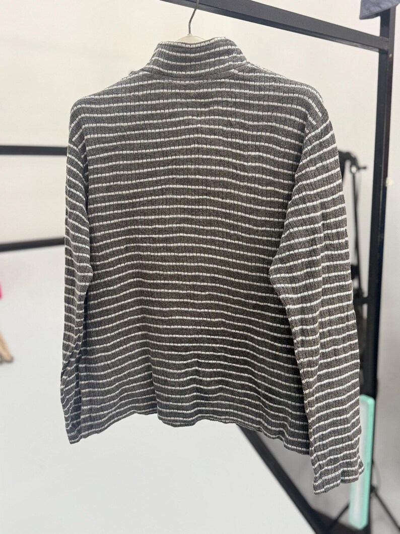 May include: A long-sleeved, ribbed knit sweater featuring a high collar and a horizontal striped pattern in dark charcoal gray and white. The garment is displayed on a hanger, showcasing the textured fabric and the consistent stripe design.