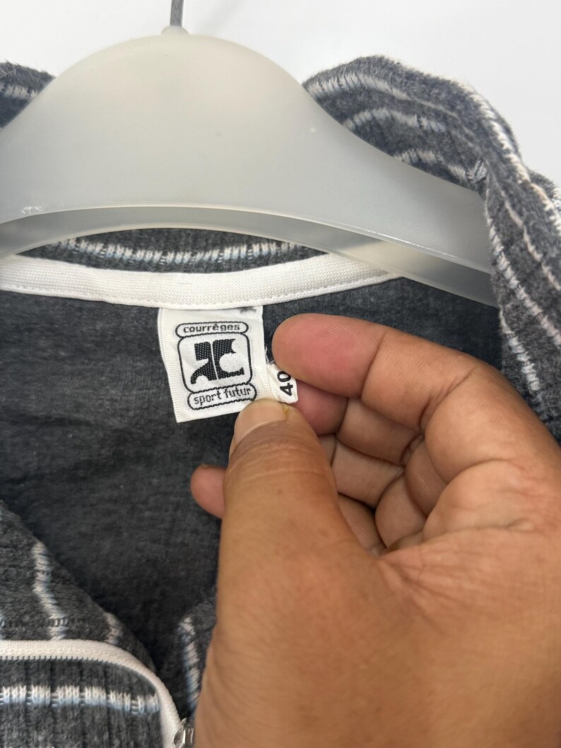 May include: A dark gray zip-up jacket with white pinstripes and a white inner collar lining. The garment features a white interior brand label with the text courr&egrave;ges sport futur and a small white size tag with the number 40 attached to the collar.