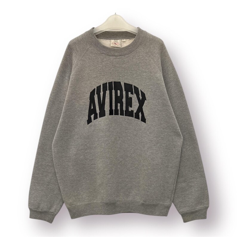 Pode incluir: Um moletom cinza mesclado com a palavra "AVIREX" impressa em letras pretas grandes e arqueadas no peito. O moletom tem gola redonda e mangas compridas. A pe&ccedil;a de roupa est&aacute; pendurada em um cabide branco.