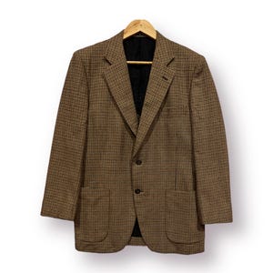 May include: A brown and tan plaid blazer with a two-button closure, two front pockets, and a black interior lining. The blazer is hanging on a wooden hanger against a white background.