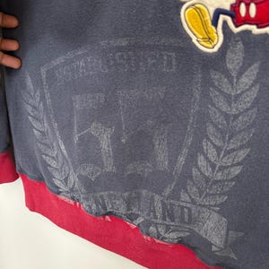 May include: Navy blue sweatshirt with a red bottom hem. Features a faded graphic of a crest with text and a Mickey Mouse patch. The sweatshirt has a vintage aesthetic.