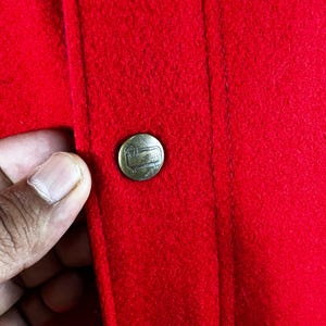 May include: A close-up of a red fabric jacket with a gold button. The button has a small logo on it.