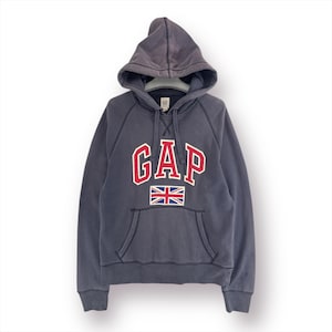 Könnte beinhalten: Ein dunkelgrauer Kapuzenpullover mit dem Wort "GAP" in roten Buchstaben mit weißem Rand. Darunter befindet sich eine kleine Union-Jack-Flagge. Der Pullover hat eine Vordertasche und eine Kapuze mit Kordelzug.