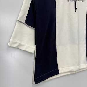 Puede incluir: Una sudadera con bloques de color en blanco y azul marino. La sudadera presenta el texto "courr&egrave;ges paris" en una fuente estilizada. Las mangas son de tres cuartos y la prenda tiene una capucha con cord&oacute;n. El dise&ntilde;o es moderno y minimalista.
