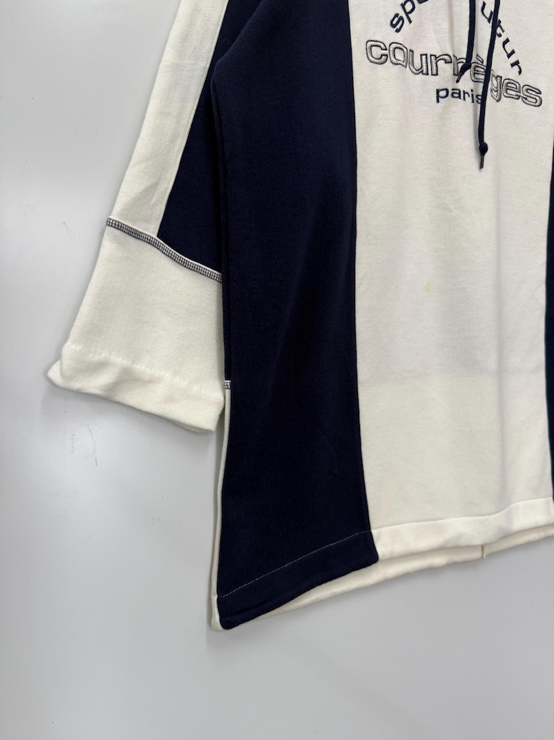 Puede incluir: Una sudadera con bloques de color en blanco y azul marino. La sudadera presenta el texto "courr&egrave;ges paris" en una fuente estilizada. Las mangas son de tres cuartos y la prenda tiene una capucha con cord&oacute;n. El dise&ntilde;o es moderno y minimalista.