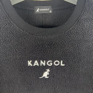 Op de afbeelding: Zwarte fleece trui met een wit geborduurd kangoeroe logo en het woord "KANGOL" op de voorkant.