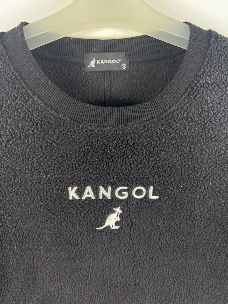 Op de afbeelding: Zwarte fleece trui met een wit geborduurd kangoeroe logo en het woord "KANGOL" op de voorkant.