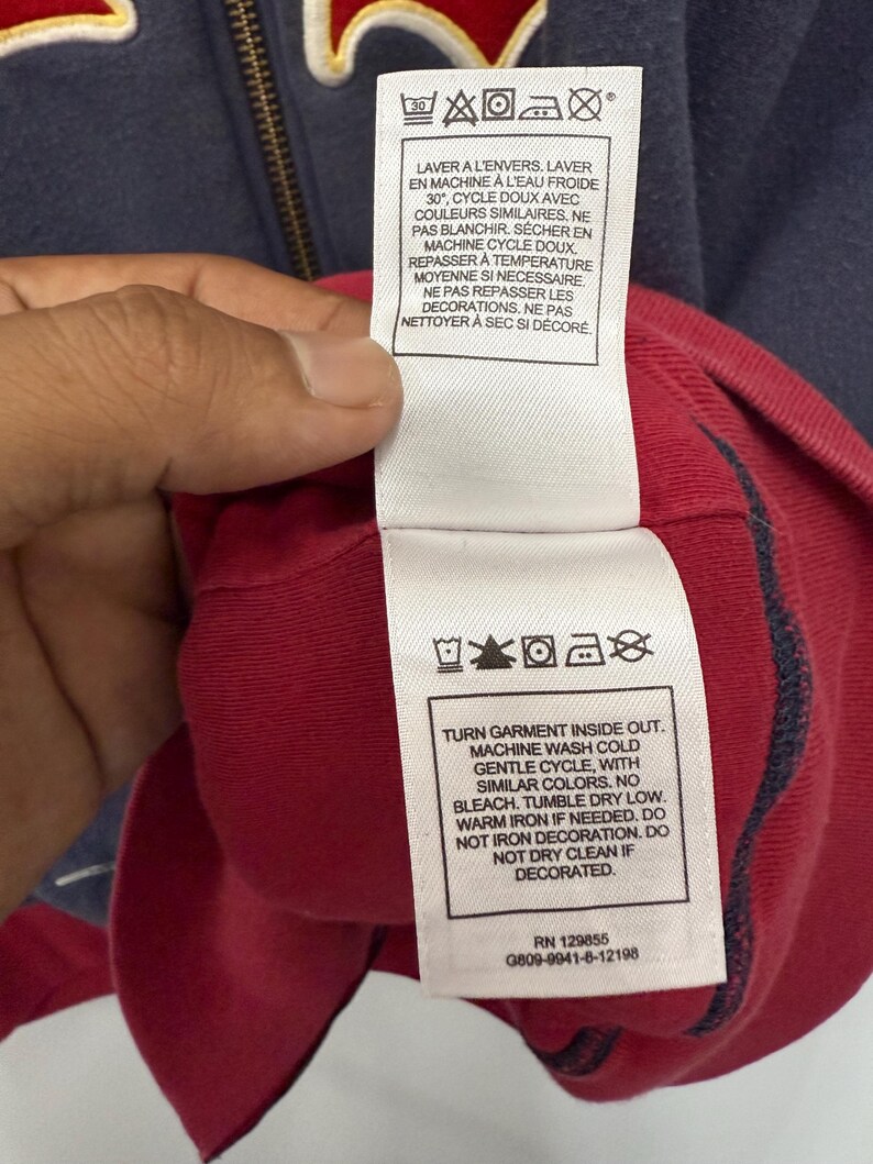 May include: Close-up of a navy blue and red garment with care instructions printed on a white tag. The tag includes washing and drying symbols, along with text in English and French. The garment has a zipper.
