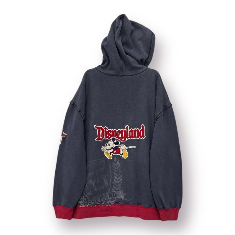May include: Navy blue hooded sweatshirt with red trim. The back features the Disneyland logo with Mickey Mouse. The sweatshirt has a vintage look and feel, with a distressed design. The sleeves have a distressed look.