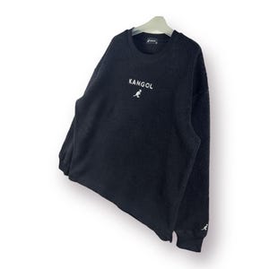 Op de afbeelding: Zwarte fleece trui met een wit kangoeroe logo en het woord "Kangol" geborduurd op de voorkant.