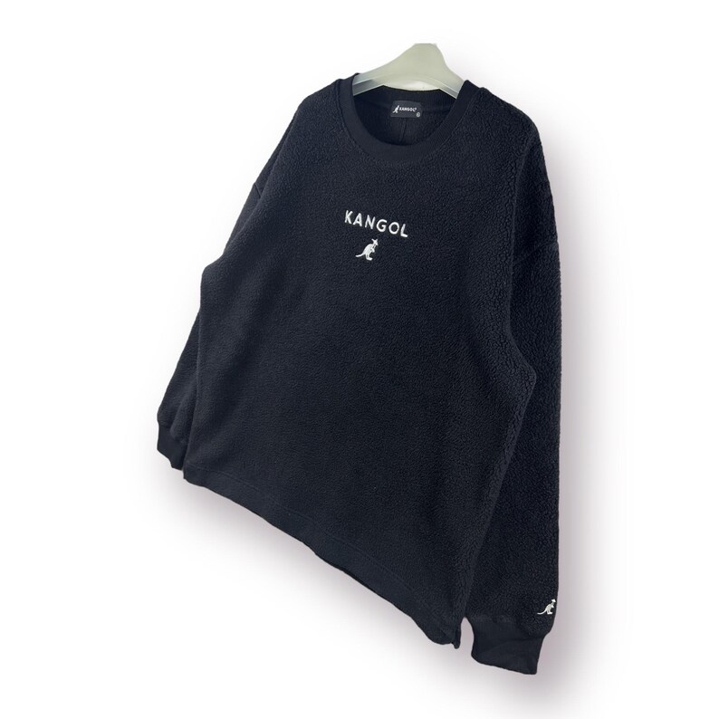 Op de afbeelding: Zwarte fleece trui met een wit kangoeroe logo en het woord "Kangol" geborduurd op de voorkant.