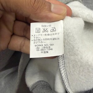 Pode incluir: Um close-up de um moletom cinza com interior branco. Uma etiqueta branca &eacute; vis&iacute;vel, com texto japon&ecirc;s e s&iacute;mbolos de cuidados. A etiqueta inclui o texto "WORKS NO.-001" e outro texto japon&ecirc;s.
