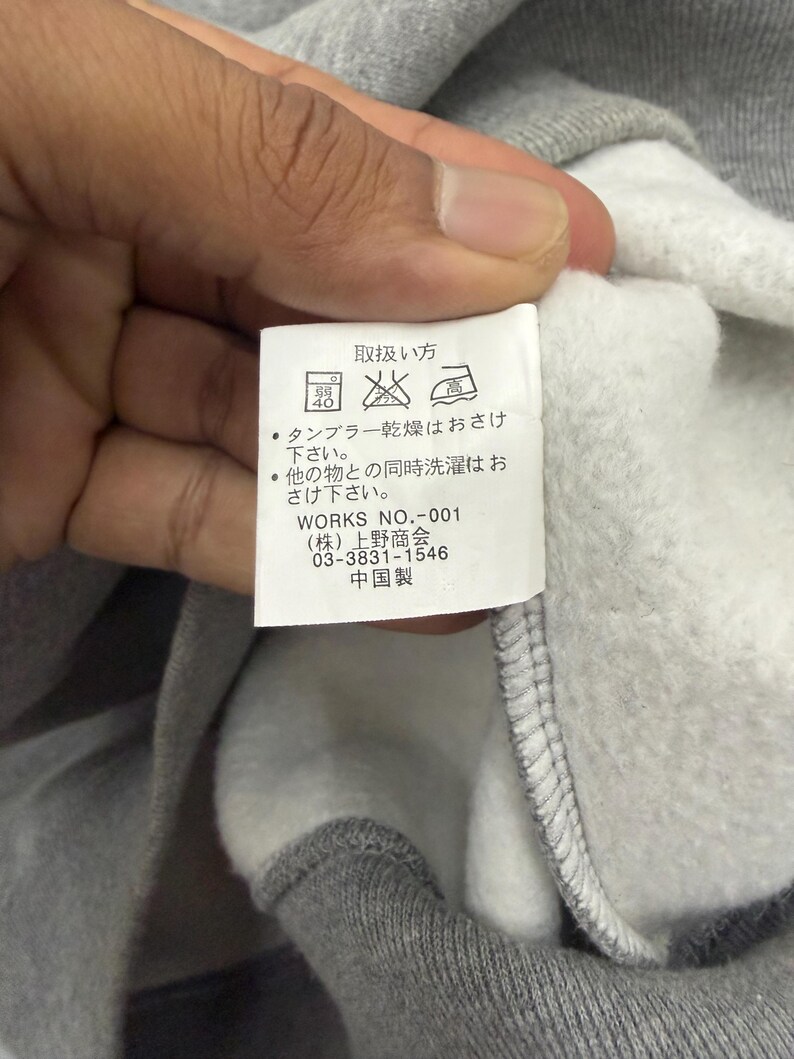 Pode incluir: Um close-up de um moletom cinza com interior branco. Uma etiqueta branca &eacute; vis&iacute;vel, com texto japon&ecirc;s e s&iacute;mbolos de cuidados. A etiqueta inclui o texto "WORKS NO.-001" e outro texto japon&ecirc;s.