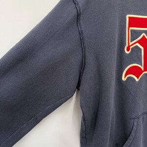 May include: A navy blue hooded sweatshirt with a red and white graphic on the front. The sweatshirt has a kangaroo pocket and a red bottom hem. The sleeves have a distressed seam.