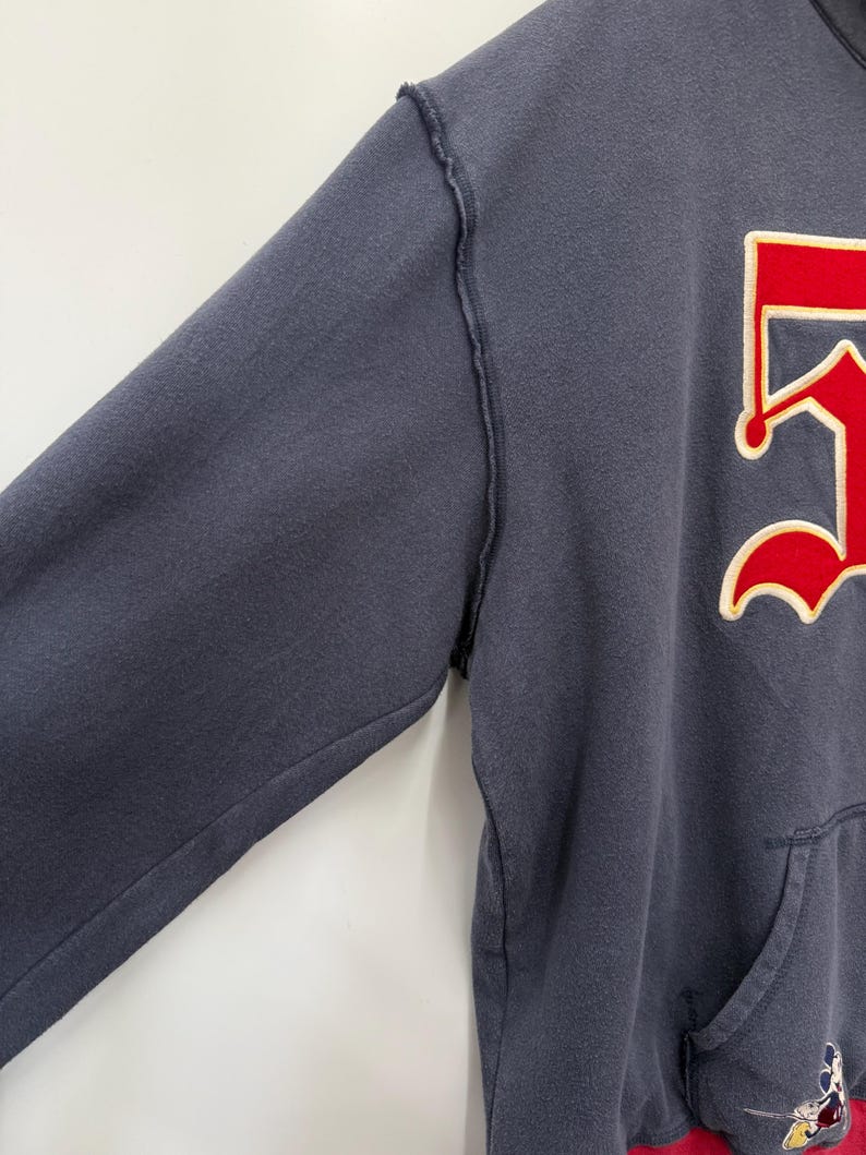 May include: A navy blue hooded sweatshirt with a red and white graphic on the front. The sweatshirt has a kangaroo pocket and a red bottom hem. The sleeves have a distressed seam.