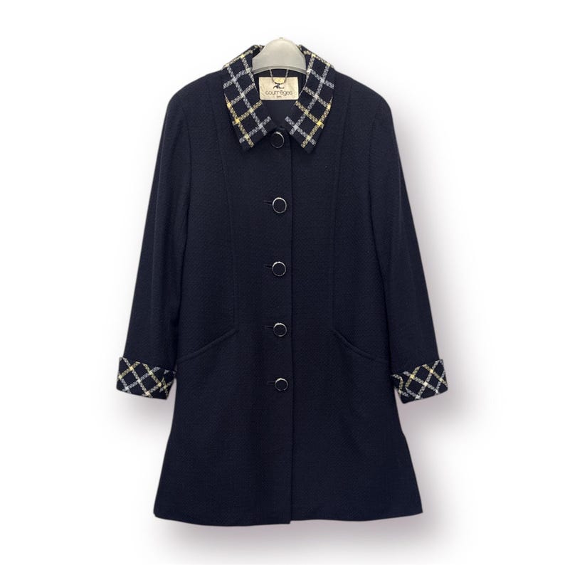 May include: A navy blue coat with a plaid collar and cuffs. The coat has a button-down front and two front pockets. The collar and cuffs feature a yellow, white, and black plaid pattern. The coat is hanging on a hanger against a white background.