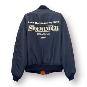May include: A navy blue bomber jacket with a white graphic on the back that says "Let's Dance in the Sky! Sidewinder PlayStation Asmik".