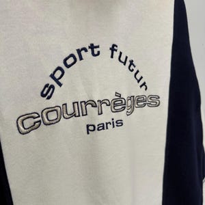 Puede incluir: Sudadera color crema con detalles en azul marino y cuello alto. La parte delantera presenta el texto bordado "sport futur" sobre "COURR&Egrave;GES paris" en hilo azul marino y plateado.