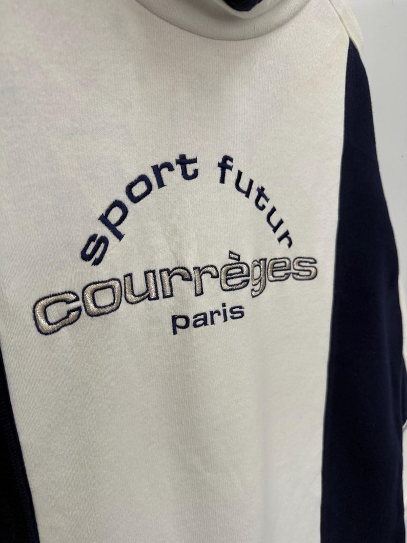Puede incluir: Sudadera color crema con detalles en azul marino y cuello alto. La parte delantera presenta el texto bordado "sport futur" sobre "COURR&Egrave;GES paris" en hilo azul marino y plateado.