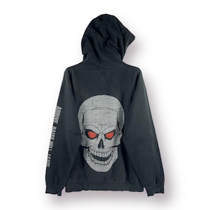 Puede incluir: Sudadera con capucha negra con un gran gráfico de calavera blanca con ojos rojos en la espalda. El texto "ARRIVE RAISE HELL LEAVE" está impreso en la manga izquierda.