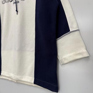 Puede incluir: Una sudadera con capucha blanca y azul marino con el texto "sport futur courr&egrave;ges paris". La sudadera tiene un dise&ntilde;o de bloques de color con un panel central blanco y paneles laterales azul marino. Las mangas son de tres cuartos.