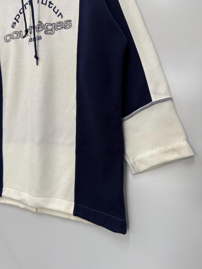 Puede incluir: Una sudadera con capucha blanca y azul marino con el texto "sport futur courr&egrave;ges paris". La sudadera tiene un dise&ntilde;o de bloques de color con un panel central blanco y paneles laterales azul marino. Las mangas son de tres cuartos.