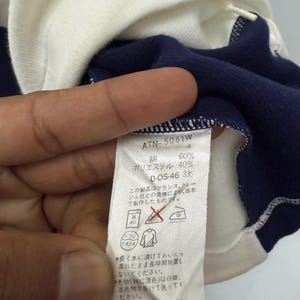 Puede incluir: Primer plano de una camiseta de manga larga azul marino y blanca. La camisa tiene cuello y pu&ntilde;os blancos. Una etiqueta con texto japon&eacute;s es visible, junto con detalles de la composici&oacute;n de la tela.
