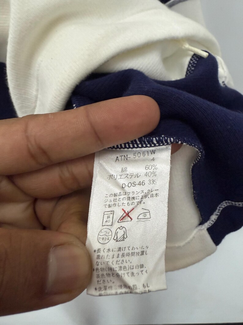 Puede incluir: Primer plano de una camiseta de manga larga azul marino y blanca. La camisa tiene cuello y pu&ntilde;os blancos. Una etiqueta con texto japon&eacute;s es visible, junto con detalles de la composici&oacute;n de la tela.