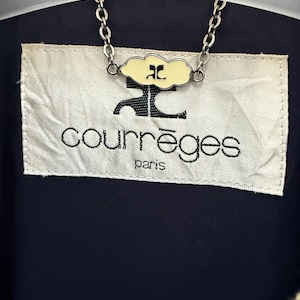 May include: Close-up of a navy blue garment with a cream-colored label. The label features the brand name "courr&egrave;ges" in black lettering, with "paris" below. A chain with a cloud-shaped charm is attached to the garment.