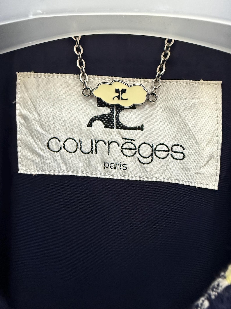 May include: Close-up of a navy blue garment with a cream-colored label. The label features the brand name "courr&egrave;ges" in black lettering, with "paris" below. A chain with a cloud-shaped charm is attached to the garment.