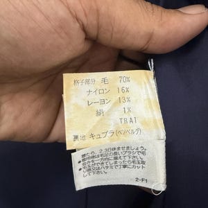 May include: A close-up of a fabric tag with Japanese text and percentages for wool, nylon, rayon, and silk. The tag is attached to a dark blue garment. Additional Japanese text provides care instructions.