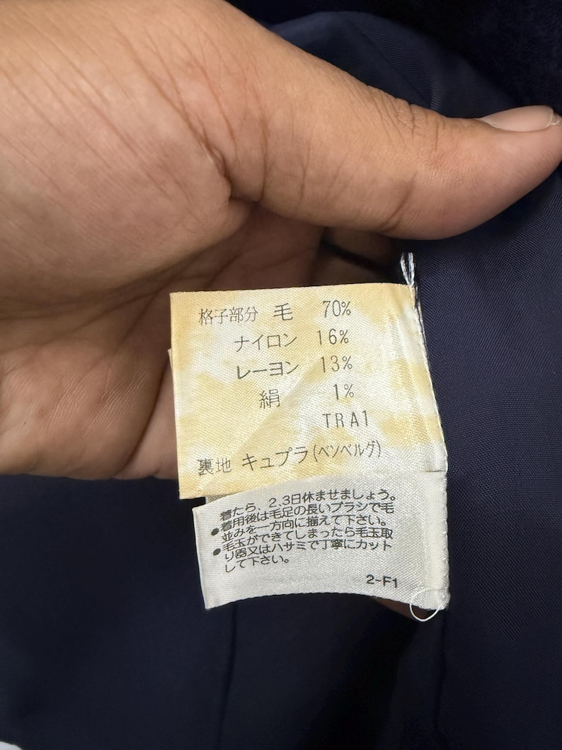 May include: A close-up of a fabric tag with Japanese text and percentages for wool, nylon, rayon, and silk. The tag is attached to a dark blue garment. Additional Japanese text provides care instructions.