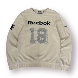 May include: Cream-colored Reebok sweatshirt with the number "18" in a gray, 3D-style font on the front. The Reebok logo is in navy blue across the chest. A small navy blue graphic is on the left sleeve.