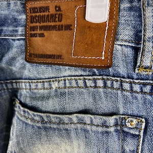 Może przedstawiać: Zbliżenie na parę niebieskich dżinsów z brązową skórzaną naszywką z napisem "Exclusive CA. DSQUARED2 Ruff Workwear Inc. Since 1995 It's a Hard Knock Life..."